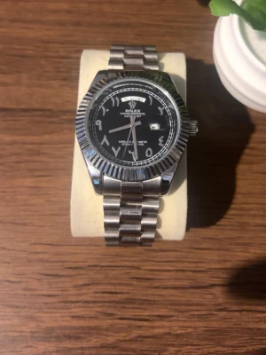 Klocka  - Klocka med svart urtavla och arabiska siffror. Klockan har ett klassiskt armband i silverfärgat stål och räfflad bezel. Perfekt accessoar för dig som gillar exklusiv och tidlös design. Priset kan diskuteras!