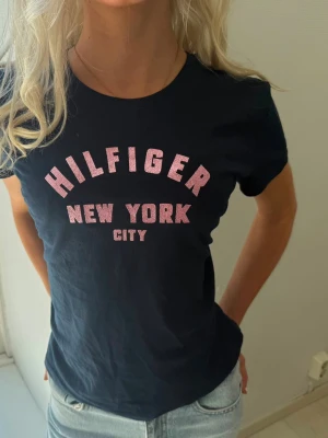 Svart Hilfiger t-shirt med tryck - Snygg svart t-shirt från Hilfiger med rosa texttryck 'HILFIGER NEW YORK CITY' på bröstet. Klassisk rund halsringning och korta ärmar. Perfekt till jeans eller shorts för en avslappnad look.