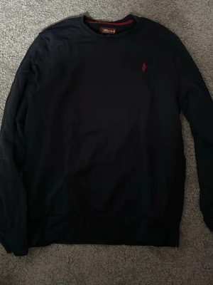 Svart sweatshirt från MCS - Klassisk svart sweatshirt från MCS med rund halsringning och diskret röd logga broderad på bröstet. Tröjan har långa ärmar och ribbade muddar vid ärmslut och nederkant. Perfekt för en avslappnad och stilren look.