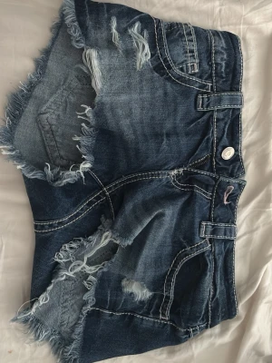 Blå slitna jeansshorts med nitar - Säljer ett par blå jeansshorts med råa, fransiga kanter och slitningar framtill. Shortsen har dekorativa nitar på bakfickorna och tydliga vita sömmar. 