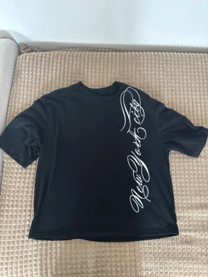Svart t-shirt med New York-tryck - Cool svart t-shirt från SUMWON i storlek XS med vit text 'New York City' i snirkligt tryck längs framsidan. T-shirten har klassisk passform och rund hals. Perfekt för dig som gillar streetstyle och vill sticka ut med en snygg detalj.