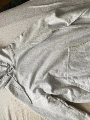 Ljusgrå hoodie med magficka - En basic ljusgrå hoodie med huva och dragsnören. Hoodien har en stor magficka framtill och är gjord i mjukt sweatshirtmaterial, troligen bomullsblandning. Perfekt för en avslappnad och chill stil. Det är en liten deo fläck på den
