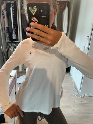 Vit långärmad topp från Ralph Lauren - Klassisk vit långärmad topp från Ralph Lauren med den ikoniska lilla loggan broderad på bröstet. Toppen har en normal passform och är tillverkad i mjuk bomull, perfekt för en clean och enkel look.