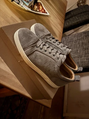 Grå loubs - Snygga loubs i grå mocka med den klassiska röda sulan och matchande grå skosnören. Klassisk låg modell med diskret design och beige innerfoder. Perfekta för dig som gillar stilrena och enkla skor till vardagslooken.