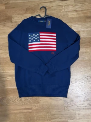 Blå stickad tröja Polo Ralph Lauren - Snygg blå stickad tröja från Polo Ralph Lauren med amerikansk flagga på bröstet och RL-broderi. Tröjan har rund halsringning, ribbstickade muddar och nederkant. Perfekt för dig som vill ha en klassisk och trendig look.