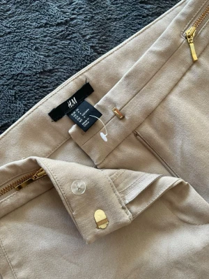 Beige kostymbyxor från H&M - Stilrena beige kostymbyxor från H&M med raka ben och två dekorativa guldfärgade dragkedjor vid fickorna. Byxorna har en klassisk passform och är tillverkade i ett mjukt, vävt material som ger en snygg och dressad look.