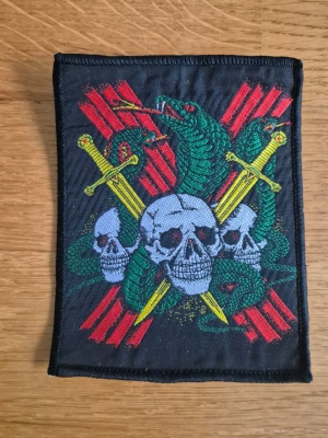 Tygmärke med dödskallar och ormar - VINTAGE Patch 1980-tal, BIKER TRIPLE SKULL, SNAKES & SWORDS Detta är ett broderat tygmärke med motiv av dödskallar, ormar och korsade svärd.Liknande motiv förekommer på vintage-bandanas från 80-talet och tygmärken för MC-västar m.m - Vintage  - Ny/oanvänd  - 13x10cm - 1980-tal  - Made in USA - MC - Patch - Tygmärke   Läs mer här --> https://share.google/SqXR64bGhAH7ALaW4  #Vintage  #patch #tygmärke  #1980  #USA #MC #skulls #swords #snakes