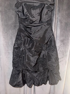 Svart axelbandslös kortklänning - Säljer en svart axelbandslös kortklänning med snörning i ryggen och draperade detaljer. Klänningen har blommiga detaljer och är även väldigt luftig 
