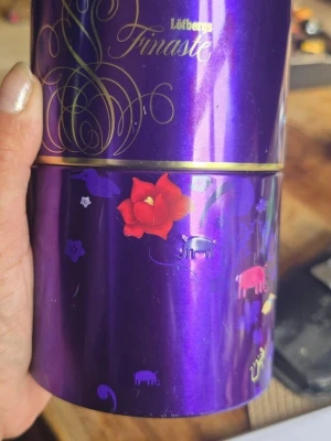 Lila Löfbergs plåtburk - Säljer en lila plåtburk från Löfbergs Finaste med gulddetaljer och färgglada motiv av blommor och djur. Perfekt för förvaring av kaffe, te eller småprylar. Burken har ett lock och är dekorerad med en stor röd blomma och små figurer. Höjd 15 cm