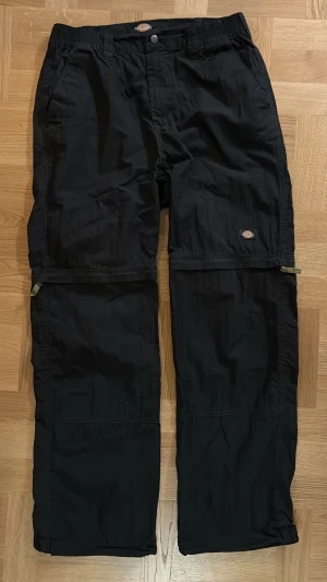 Svarta zip-off byxor från Dickies - Svarta byxor från Dickies med zip-off funktion så du kan göra om dem till shorts. Byxorna har raka ben, resår i midjan, bälteshällor och flera fickor både fram och bak. Tillverkade i slitstarkt syntetmaterial, perfekta för en aktiv stil.