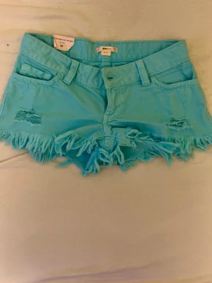 Turkosa shorts från gina tricot - Säljer ett par turkosa shorts från gina med fransig kant och slitna detaljer framtill.