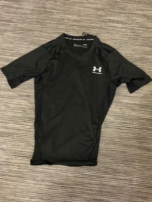 Svart träningströja Under Armour - Säljer en svart, tight och kortärmad träningströja från Under Armour. Tröjan har rund halsringning och logga i vitt på bröstet. Tillverkad i ett snabbtorkande, stretchigt funktionsmaterial som passar perfekt till gym eller löpning.