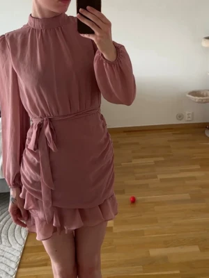 Rosa omlottklänning med volang - Supersöt kort rosa klänning med långa ärmar, volangkant och omlottdetalj i midjan. Klänningen har ett knytband i midjan och lätt rynkade detaljer längs sidorna för en snygg siluett. Perfekt för dig som gillar romantisk och trendig stil.