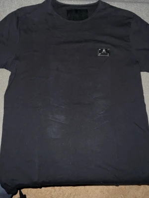 Svart Philipp Plein t-shirt med patch - Säljer en svart t-shirt från Philipp Plein med en snygg metallpatch på bröstet med döskalle och logga. T-shirten har rund hals och korta ärmar, perfekt för en stilren och edgy look.