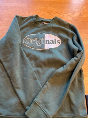Grön sweatshirt från Jack & Jones - Mysig grön sweatshirt från Jack & Jones med stort 'Originals'-tryck på bröstet i vitt och svart. Tröjan har rund halsringning och ribbade muddar vid ärmslut och nederkant. Perfekt för en avslappnad stil. Aldrig använd.