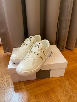 Vita Valentino sneakers med guld nitar - Snygga vita sneakers från Valentino med guldfärgade nitar på ovansidan och klassisk låg profil. Skorna har en stilren design med vita snören och detaljer i läder. Perfekta för dig som vill ha en unik touch på din outfit. Allt medföljer och det finns flera storlekar