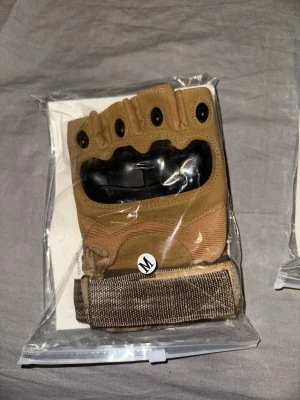Bruna halvfingerhandskar med skydd - Säljer ett par bruna halvfingerhandskar med förstärkt knogskydd i svart plast och justerbart kardborreband vid handleden. Perfekta för träning eller utomhusaktiviteter där du vill ha extra grepp och skydd. Storlek M.