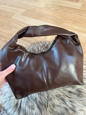 Mörkbrun elegant handväska  - Säljer en snygg mörkbrun handväska i mjukt läder med bredt handtag och minimalistisk design. Väskan har en slät yta och synliga sömmar framtill. Perfekt för dig som gillar stilrena accessoarer.