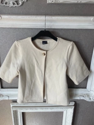 Kortärmad beige kofta från Gina Tricot - Stilren beige kofta från Gina Tricot med korta ärmar och rund halsringning. Koftan har en enkel knäppning framtill med en dekorativ guldig knapp.