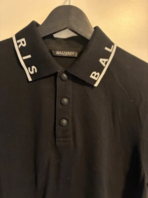 Svart pikétröja från Balmain - Snygg svart pikétröja från Balmain med vit text på kragen där det står 'PARIS BAL'. Tröjan har korta ärmar och knappar framtill. Klassisk och stilren design med vit detalj på ärmslutet. Perfekt för en trendig och avslappnad look.