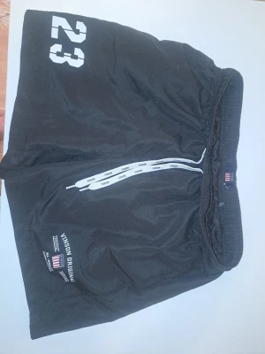 Svarta shorts från Vinson Original - Säljer ett par svarta shorts från Vinson Original med vitt tryck av siffran 23 på vänster ben och logga på höger ben. Shortsen har resår i midjan och vita dragsnören med text. Perfekta för en sportig look.