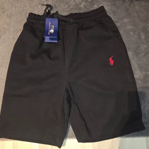 Svarta shorts från Polo Ralph Lauren - Svarta shorts från Polo Ralph Lauren med elastisk midja och snörning. Klassisk passform med två sidofickor och en liten röd logga broderad på vänster ben. Perfekta för sommaren och avslappnade dagar.