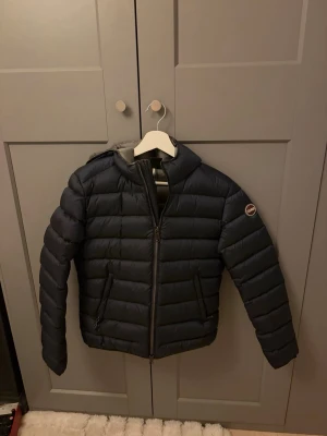 Mörkblå Colmar jacka - Snygg mörkblå pufferjacka med huva och dragkedja framtill. Jackan har två sidofickor med dragkedja och ett märke på ena ärmen. Quiltad design med horisontella sömmar som ger en klassisk pufferlook. Perfekt för kalla vinterdagar. Slimfit, mörklblå och ny 