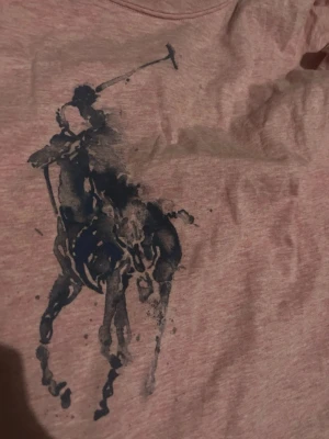 Rosa t-shirt med blått Polo-tryck - Säljer en rosa t-shirt från Polo Ralph Lauren med det klassiska blå polospelar-trycket framtill. T-shirten har korta ärmar och är gjord i mjuk bomull, perfekt för en avslappnad och stilren look.