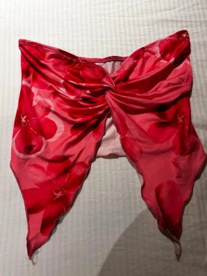 Blommig bandeau tubtopp i rosa och rött - Trendig bandeau tubtopp med stora blommor i rosa och rött. Toppen har en snygg twist-detalj framtill och spetsiga kanter nedtill. Tillverkad i ett tunt, stretchigt syntetmaterial som känns lätt och somrig.