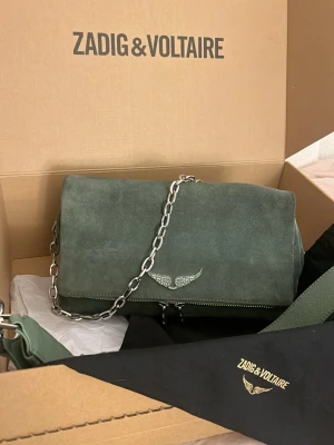 Zadig väska - Så fin zadigväska i grön mocka med silvrig kedja, använd ett fåtal gånger, skicket superfint. Inga defekter, rymmer väldigt mycket. Köpt helt ny, allt som man får med när man köper den ny tillkommer, dustbag, ett längre axelband och originalkartongen. Lika väska säljs begagnad för runt 4700kr. Kommer tyvärr inte ihåg nypris men vet att den låg på över 5000, väskan säljs inte längre ny men denna är i nyskick. Säljer då den tyvärr inte kommer till användning och vill se vad jag kan få för den.