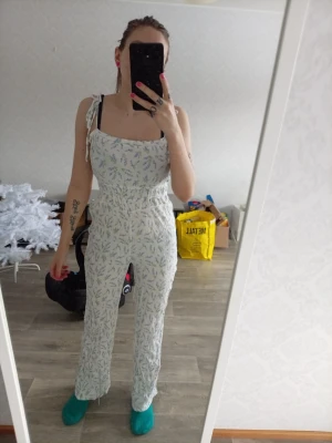 Vit blommig jumpsuit med knytband - Supersöt vit jumpsuit med små blommor i grönt och blått. Har knytband på axlarna och resår i midjan för skön passform. Vida ben och mjukt, lätt tyg som känns luftigt. Perfekt för dig som gillar en avslappnad men ändå trendig stil.