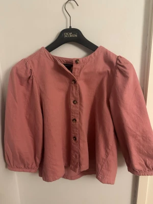 Rosa blus med knappar från Lindex - Söt rosa blus från Lindex med trekvartsärm och dekorativa bruna knappar framtill. Blusen har en rund halsringning och lätt puffade axlar för en trendig look. Tillverkad i ett mjukt tyg som känns bekvämt mot huden. aldrig använd!
