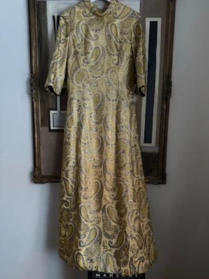 Guld paisleymönstrad långklänning vintage festklänning  - Unik guld långklänning med paisleymönster i guld och blå inslag.. Klänningen har hög krage, halvlånga ärmar och en elegant, utsvängd siluett. Materialet känns lyxigt och skimrande, perfekt för dig som vill sticka ut. Storleken uppskattas till en 40/42