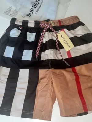 Burberry badbyxor med rutigt mönster - Snygga badbyxor från Burberry med klassiskt rutigt mönster i svart, beige, vitt och rött. De har en elastisk midja med dragsko i rött och vitt samt Burberry-tag. Perfekta för stranden eller poolen och ger en exklusiv känsla.