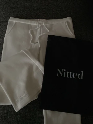 Low bootcut mjukisbyxor - Säljer ett par vita mjukisbyxor från Nitted med broderad logga vid benet. Byxorna har låg midja och utsvängda ben, sidofickor och snören i midjan. Tillverkade i mjukt material och är både bekväma och stilrena.