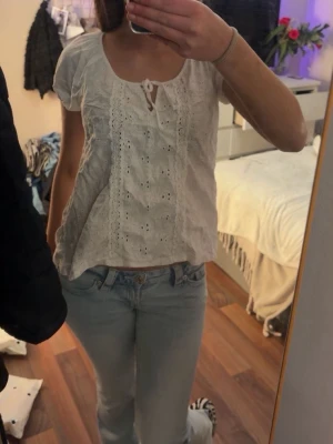 Vit blus med spetsdetaljer och knyt - Söt vit blus med korta ärmar, spetsdetaljer framtill och knyt vid halsen. Blusen har broderade hålmönster och en luftig passform som ger en romantisk vibe. Perfekt till jeans eller kjol för en fräsch look.