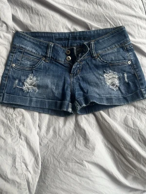 lågmidjade Blå jeansshorts med slitningar - Säljer ett par blå denimshorts  med slitna detaljer framtill och klassiska fem fickor. Shortsen har dubbla knappar i midjan och en läderpatch bak. Perfekta för sommaren och ger en avslappnad look. köpta för 500 därav pris