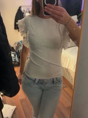 Vit topp med volangärmar - Säljer en vit topp med volangdetaljer på axlarna och korta ärmar. Toppen har en rund halsringning och är figurnära i modellen. Perfekt för dig som vill ha något stilrent men ändå med en twist.