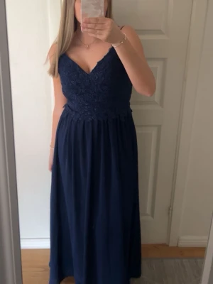 Marinblå långklänning med spetsliv - Säljer en marinblå långklänning med smala axelband och v-ringad hals. Livet är dekorerat med spets och har fina broderade detaljer. Kjolen faller mjukt och klänningen har en elegant och tidlös look. Perfekt för dig som vill sticka ut på bal eller fest.