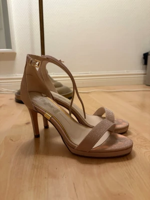 Glittriga nude pumps med ankelrem - Säljer ett par snygga pumps i nude med glittrig finish. Skorna har öppen tå, smal ankelrem med guldfärgat spänne och en elegant klack. Perfekta för att lyfta din outfit med lite extra glam. Materialet är textil med glittereffekt.