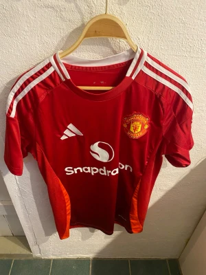 Manchester United matchtröja röd - Säljer en röd Manchester United matchtröja från Adidas med vita och röda detaljer, klubbmärke på bröstet och Snapdragon som sponsortryck. Tröjan har korta ärmar, rund hals och är tillverkad i ett lätt, ventilerande funktionsmaterial.