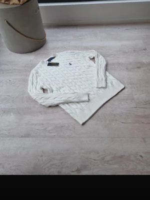 Vit kabelstickad tröja Polo Ralph Lauren - Kabelstickad vit tröja från Polo Ralph Lauren med rund halsringning och diskret broderad logga på bröstet. Tröjan har långa ärmar och ribbade muddar vid ärmslut och nederkant. Klassisk och tidlös design i mjukt material.