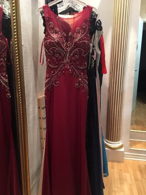 Vinröd långklänning med paljetter - Vinröd balklänning med transparent överdel och  paljetter. Klänningen är ärmlös och har en markerad midja. Använd bara en gång på min egen bal.