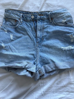 Blå denimshorts från h&m - Säljer ett par ljusblå denimshorts från h&m med hög midja och avslappnad passform. Shortsen har slitna detaljer, uppvikta benslut och klassiska fem fickor. Perfekta för sommaren och ger en cool, avslappnad vibe.