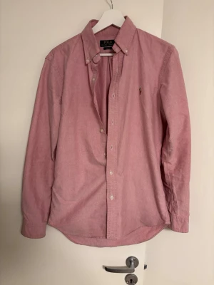 Rosa skjorta från Polo Ralph Lauren - Klassisk rosa skjorta från Polo Ralph Lauren i slim fit. Skjortan har knappar framtill, button-down krage och den ikoniska broderade loggan på bröstet. Tillverkad i mjuk bomull och har långa ärmar. Perfekt för en clean och stilren look.