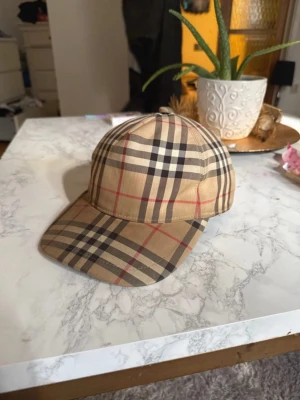 Beige keps från Burberry med rutmönster - Säljer en klassisk beige keps från Burberry med det ikoniska rutmönstret i svart, vitt och rött. Kepsen har böjd skärm och justerbar rem bak. Tillverkad i bomullsmix och har en stilren look som passar till många outfits.