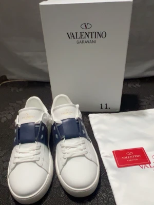 Valentino Garavani  - Säljer ett par snygga Valentino Garavani sneakers i vitt med marinblå detalj över snörningen. Skorna har rund tå, platt sula och är tillverkade i skinn. Klassisk design med vita snören och diskret logga på plösen. Kommer med originalkartong och dustbag.