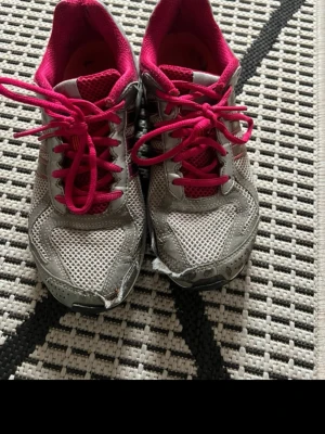 Grå och rosa sneakers från Adidas - Snygga sneakers från Adidas i grått meshmaterial med rosa detaljer och skosnören. Låg modell med ventilerande ovandel och bekväm innersula. Perfekta för dig som gillar sportig stil och vill ha ett par lätta skor till vardagen.