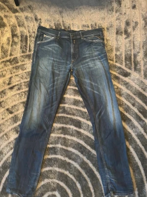 Blå slim fit jeans med slitningar - Säljer ett par blå replay, slim fit jeans med snygga slitningar och tvättade detaljer. Klassisk femficksmodell med dragkedja och knapp i midjan. Jeansen har kontrastsömmar och raka ben. Perfekta för en avslappnad och trendig look.