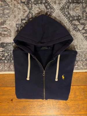 Mörkblå Polo Ralph Lauren Zip Up Hoodie - Hej!👋  Säljer en mörkblå zip up hoodie från Polo Ralph Lauren. Hoodien har två små fläckar (se de två sista bilderna), men det är inget som riktigt märks vid användning. Har haft den länge och har aldrig ens lagt märkte till dem. Storlek: M Mått skickas privat vid begäran!😀✅ Skickar snabbt!🚛📦 Hör av er vid övriga frågor!💯